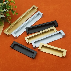 May include: An assortment of rectangular cabinet pulls in various metallic finishes, including gold, silver, and black. The pulls are arranged on an orange surface, showcasing their different colors and designs. The pulls are made of metal.