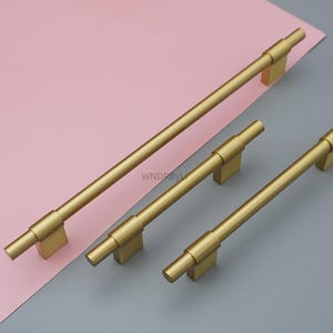 Op de afbeelding: Drie goudkleurige kastgrepen van verschillende lengtes. De grepen hebben een cilindrisch staafontwerp met vierkante montagepalen. De langste greep is ongeveer 30 cm lang. De achtergrond is een combinatie van roze en grijs.