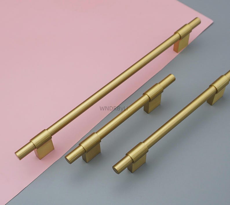 Op de afbeelding: Drie goudkleurige kastgrepen van verschillende lengtes. De grepen hebben een cilindrisch staafontwerp met vierkante montagepalen. De langste greep is ongeveer 30 cm lang. De achtergrond is een combinatie van roze en grijs.