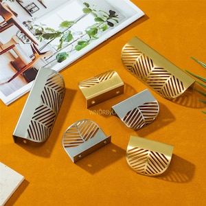 Può includere: Un assortimento di pomelli decorativi in metallo dorato e argentato. I pomelli presentano un design a forma di foglia con motivi ritagliati. L'hardware è disposto su una superficie arancione, con una rivista e del verde sullo sfondo.