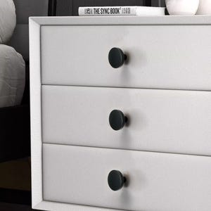 May include: A white dresser with three drawers, each featuring a black, round knob. The knobs are evenly spaced and provide a minimalist aesthetic. The top of the dresser has a book titled "THE SYNC BOOK".