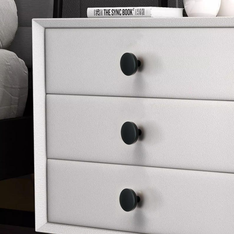 May include: A white dresser with three drawers, each featuring a black, round knob. The knobs are evenly spaced and provide a minimalist aesthetic. The top of the dresser has a book titled "THE SYNC BOOK".