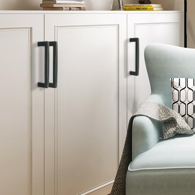 May include: A white cabinet with dark bronze rectangular handles. The handles are mounted vertically on the cabinet doors. A light blue armchair with a patterned cushion is in the background.