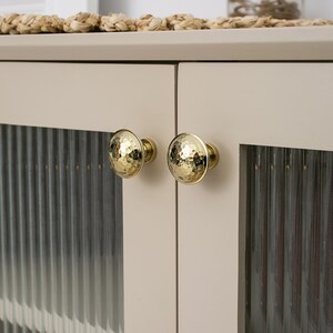 May include: Close-up of a cabinet with two gold hammered-look knobs. The cabinet doors are a light beige color, with a ribbed glass panel. The knobs are round and have a textured surface. The cabinet is part of a larger furniture piece.