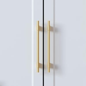 Op de afbeelding: Close-up van twee goudkleurige kastgrepen tegen een witte kastdeur. De grepen zijn cilindrisch met een verticaal staafontwerp. De kastdeuren zijn wit met een verticale zwarte lijn ertussen.