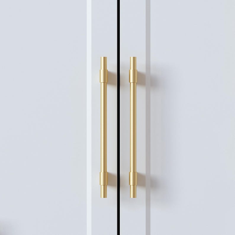 Op de afbeelding: Close-up van twee goudkleurige kastgrepen tegen een witte kastdeur. De grepen zijn cilindrisch met een verticaal staafontwerp. De kastdeuren zijn wit met een verticale zwarte lijn ertussen.