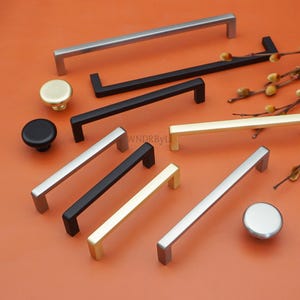 May include: An assortment of cabinet hardware, including silver, black, and gold-toned handles and knobs. The handles are rectangular, and the knobs are round. The hardware is displayed on an orange background.