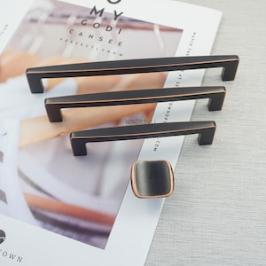 May include: A set of four bronze-coloured cabinet hardware pieces. The set includes three rectangular bar pulls of varying lengths and a square knob. The hardware is displayed on a white and grey background.