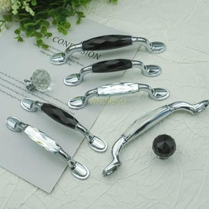 May include: A collection of cabinet hardware, including silver-toned handles with faceted black and clear crystal accents. A clear crystal knob and a faceted black knob are also present. The hardware is displayed on a light background.