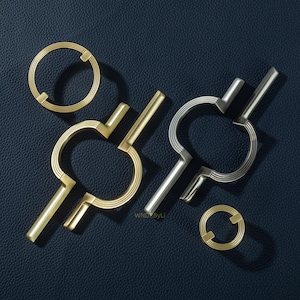 Puede incluir: Un surtido de tiradores y pomos dorados y plateados para armarios. Los herrajes presentan un diseño circular con detalles lineales y acentos angulares. Los tiradores y pomos están dispuestos sobre una superficie azul oscuro.