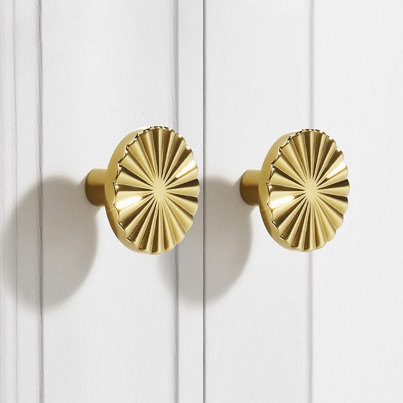 May include: Two gold-colored cabinet knobs with a sunburst design. The knobs are round with a ridged, fan-like pattern radiating from the center. They are mounted on a white surface.