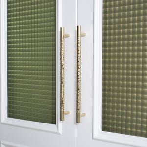May include: Close-up of a white door with two gold-colored, textured door handles. The handles are long and cylindrical, with a detailed pattern. The door has a green, textured glass panel.