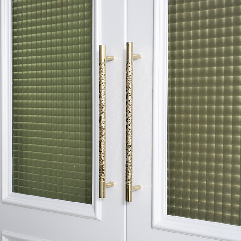 May include: Close-up of a white door with two gold-colored, textured door handles. The handles are long and cylindrical, with a detailed pattern. The door has a green, textured glass panel.