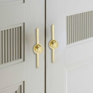 May include: Close-up of two cabinet doors with gold-coloured hardware. Each door features a vertical gold pull with a circular knob. The doors are painted a light grey colour and have a slatted design.