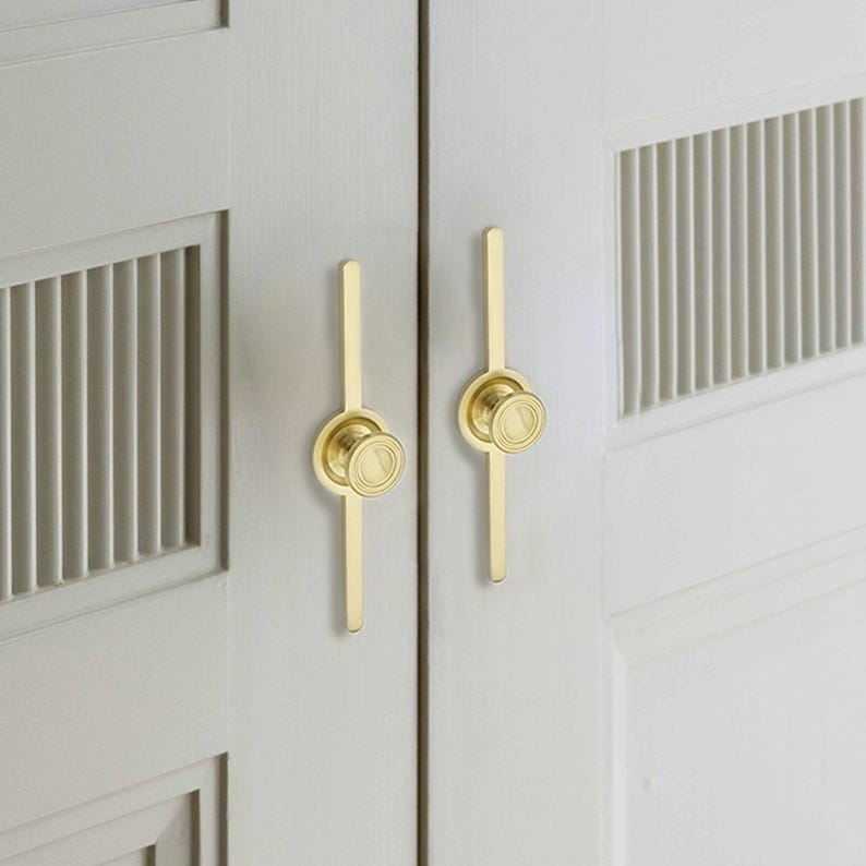 May include: Close-up of two cabinet doors with gold-coloured hardware. Each door features a vertical gold pull with a circular knob. The doors are painted a light grey colour and have a slatted design.