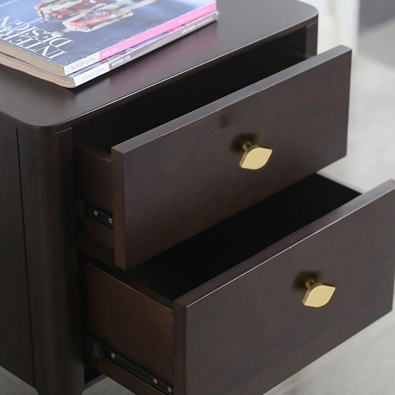 May include: Dark brown wooden bedside table with two open drawers. The drawers have gold leaf-shaped handles. A magazine rests on top of the table.