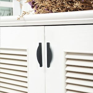 May include: A close-up of a white cabinet with two black, curved handles. The cabinet doors have a slatted design. Dried flowers and foliage are visible on the top of the cabinet.