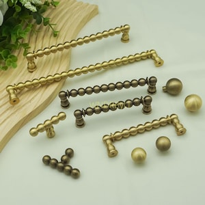 Op de afbeelding: Een verzameling messingkleurige kastbeslagen, waaronder handgrepen en knoppen. De handgrepen hebben een kralenontwerp, terwijl de knoppen bolvormig zijn. De hardware wordt op een lichte achtergrond weergegeven, met verschillende maten en stijlen voor interieurdecoratie.