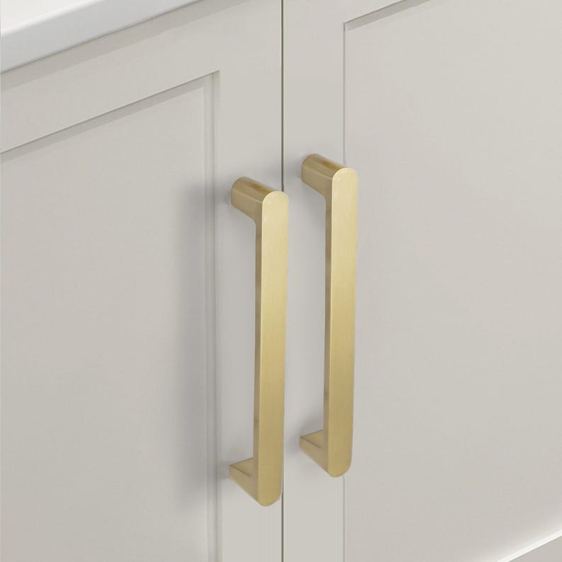 May include: Close-up of two gold-colored cabinet pulls mounted on a white cabinet door. The pulls are rectangular with rounded edges and a vertical orientation. The cabinet door has a recessed panel design.