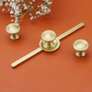 May include: A set of gold-coloured cabinet hardware, comprising two round knobs and a long pull with a central knob. The hardware features a classic design with a ridged edge. The text "WNDRByLi" is visible on the pull. White flowers are in the background.