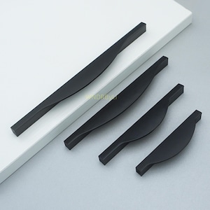 May include: Four matte black cabinet pulls in varying sizes. Each pull features a rectangular bar that twists into a curved, ergonomic handle. The pulls are arranged on a white surface, showcasing their modern design.