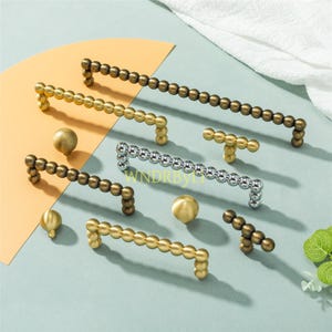 Può includere: Una collezione di ferramenta decorativa per mobili, tra cui maniglie e pomelli. Le maniglie sono in metallo e presentano un design a perline in tonalità oro, argento e bronzo. I pomelli sono sferici e abbinati.