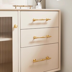 Peut inclure: Un meuble cr&egrave;me avec trois tiroirs et une porte vitr&eacute;e. Le meuble est orn&eacute; de poign&eacute;es dor&eacute;es et d'un bouton de porte rond assorti. Il s'agit d'un meuble.