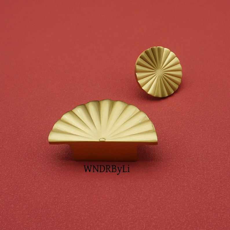 May include: Two gold-colored cabinet hardware pieces on a red background. One is a half-circle pull with a fan-like design, and the other is a round knob with a similar pattern. The text "WNDRByLi" is visible on the bottom.