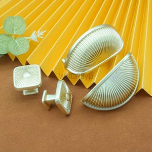 Peut inclure: Un ensemble de quincaillerie de placard dorée, comprenant deux poignées en forme de demi-lune et deux boutons carrés avec un motif de soleil. La quincaillerie est présentée sur un fond jaune en forme d'éventail et une surface marron.