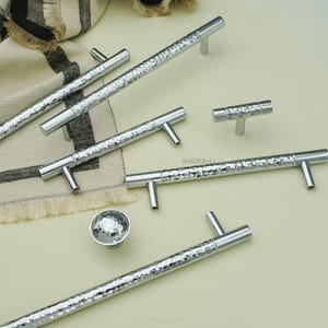May include: A collection of silver cabinet hardware, including hammered-texture bar pulls and T-shaped pulls, along with a round knob. The hardware is displayed on a light-colored surface, suggesting a modern or contemporary design aesthetic.
