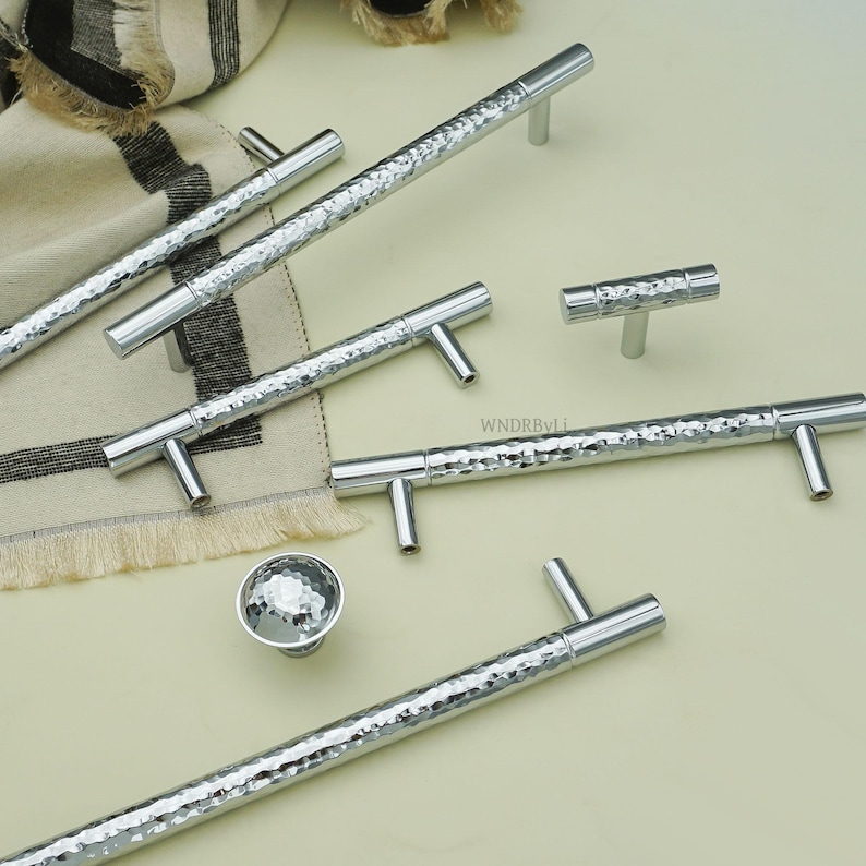May include: A collection of silver cabinet hardware, including hammered-texture bar pulls and T-shaped pulls, along with a round knob. The hardware is displayed on a light-colored surface, suggesting a modern or contemporary design aesthetic.