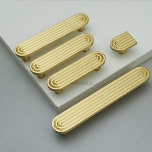 Op de afbeelding: Een verzameling goudkleurige kastbeslagen, waaronder verschillende langwerpige handgrepen en een enkele knop. De hardware heeft een gegroefd, lineair ontwerp en afgeronde uiteinden. De stukken zijn op een wit oppervlak geplaatst.