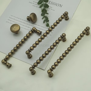 Op de afbeelding: Antieke messing kastbeslagset. De set bevat twee ronde knoppen, een kleine trekker en drie kralen trekkers. De trekkers zijn ongeveer 15 cm lang. De hardware wordt weergegeven op een wit oppervlak met tekst.