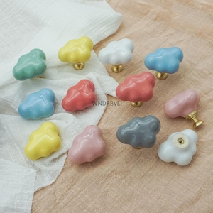 May include: A selection of cloud-shaped ceramic cabinet knobs in pastel shades of green, yellow, white, blue, red, and pink. Each knob features a brass base and a whimsical design. The knobs are displayed on a white cloth. Measurements are in cm.
