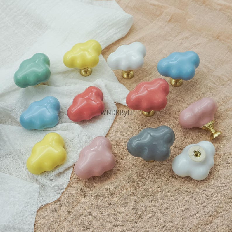 May include: A selection of cloud-shaped ceramic cabinet knobs in pastel shades of green, yellow, white, blue, red, and pink. Each knob features a brass base and a whimsical design. The knobs are displayed on a white cloth. Measurements are in cm.