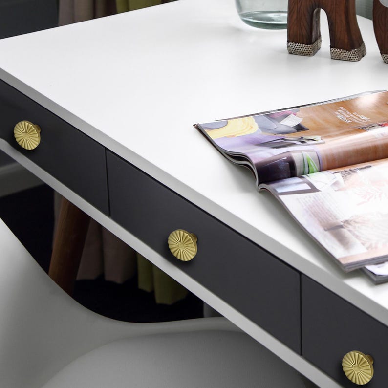 May include: A modern white desk with a black drawer section. The drawers have gold, sunburst-patterned knobs. An open magazine rests on the desk's surface. The desk is part of a contemporary home office or bedroom furniture set.