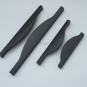 May include: Four matte black cabinet pulls in varying sizes. The pulls have a curved, ergonomic design with a slightly twisted appearance. The hardware is shown against a light gray background. The text "WNDRByLi" is visible on the image.