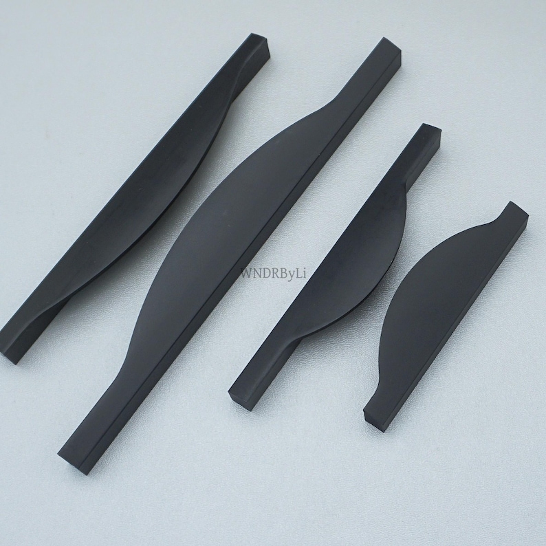 May include: Four matte black cabinet pulls in varying sizes. The pulls have a curved, ergonomic design with a slightly twisted appearance. The hardware is shown against a light gray background. The text "WNDRByLi" is visible on the image.