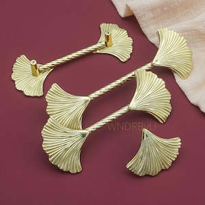 May include: Four gold-colored cabinet pulls with a ginkgo leaf design. The pulls have a textured, twisted center and a leaf-shaped end. The hardware is set against a burgundy background with a beige cloth in the upper right corner.