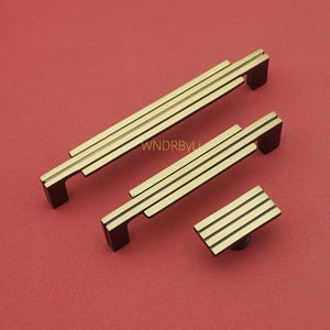 Puede incluir: Un juego de tres piezas de herrajes para armarios de color lat&oacute;n sobre un fondo rojo. El juego incluye dos tiradores rectangulares y un pomo cuadrado, todos con un dise&ntilde;o de tres barras paralelas.