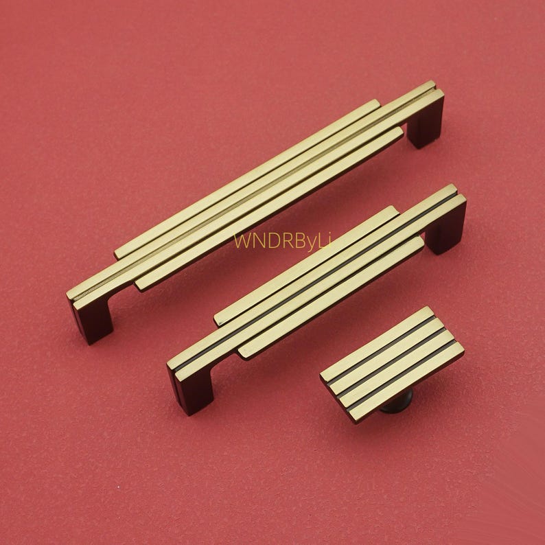 Puede incluir: Un juego de tres piezas de herrajes para armarios de color lat&oacute;n sobre un fondo rojo. El juego incluye dos tiradores rectangulares y un pomo cuadrado, todos con un dise&ntilde;o de tres barras paralelas.