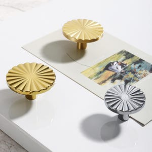 May include: Decorative cabinet knobs in gold and silver. The knobs have a fluted, sunburst design. The gold knobs are made of brass. The silver knob is made of polished metal. The knobs are on a white surface with a picture in the background.