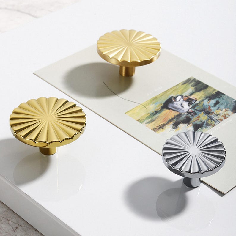 May include: Decorative cabinet knobs in gold and silver. The knobs have a fluted, sunburst design. The gold knobs are made of brass. The silver knob is made of polished metal. The knobs are on a white surface with a picture in the background.