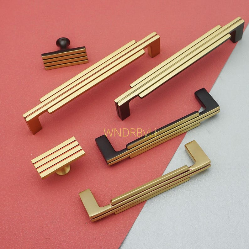 Puede incluir: Un conjunto de herrajes dorados y negros para armarios, que incluyen tiradores y pomos. Los tiradores presentan un dise&ntilde;o lineal con tres barras doradas paralelas y detalles en negro. Se exhiben sobre una superficie roja y gris.