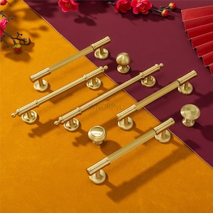 May include: A collection of gold-colored cabinet hardware, including bar pulls and round knobs. The pulls have a ribbed design, and the knobs are smooth. The hardware is arranged on a yellow and burgundy surface, with red flowers in the background.