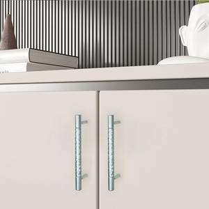 May include: Close-up of a light beige cabinet with two silver, textured handles. The cabinet is part of a larger piece of furniture, with a shelf above it holding books and decorative items. The background features vertical black and white stripes.