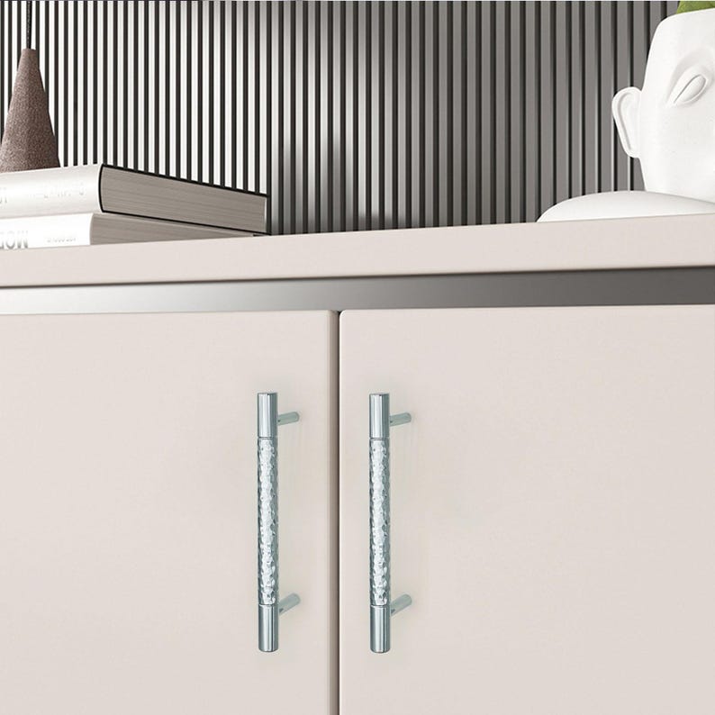 May include: Close-up of a light beige cabinet with two silver, textured handles. The cabinet is part of a larger piece of furniture, with a shelf above it holding books and decorative items. The background features vertical black and white stripes.