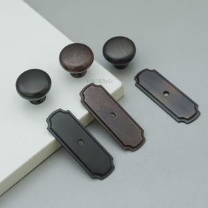 May include: An assortment of bronze and black cabinet hardware, including round knobs and rectangular backplates. The knobs have a textured finish, while the backplates feature a decorative edge. The hardware is displayed on a neutral background.