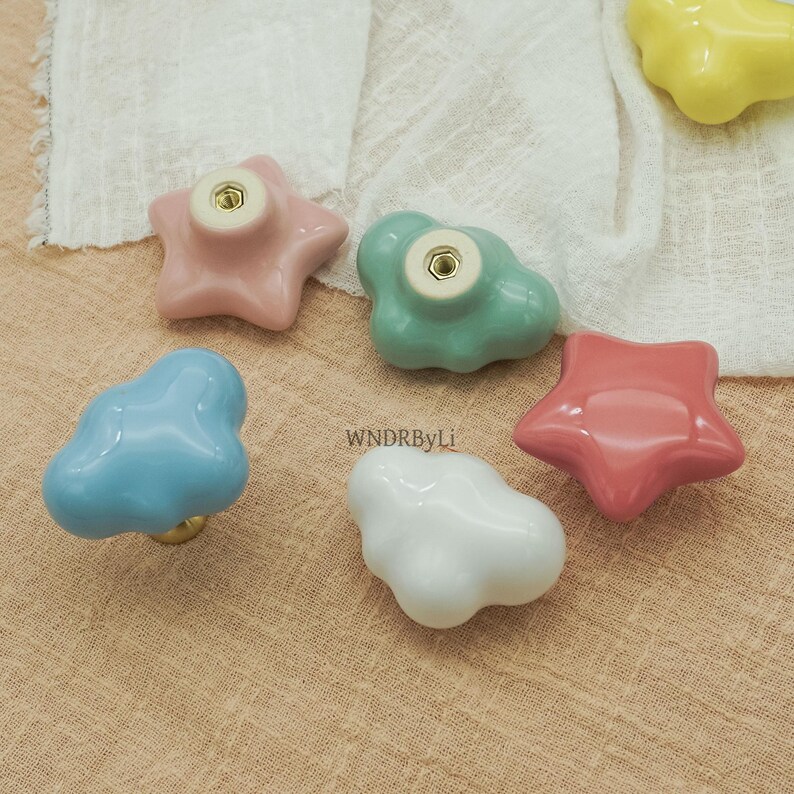 May include: A selection of colourful ceramic cabinet knobs in star and cloud shapes. The knobs are in shades of pink, blue, green, white, and yellow. Each knob has a gold-coloured screw fitting.