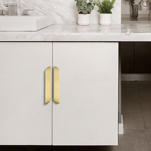 May include: A white cabinet featuring two vertical, gold-coloured handles. The handles have a curved design. The cabinet is set in a bathroom, with a white countertop and small potted plants visible in the background.